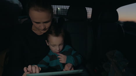 Vista-De-Una-Mujer-Con-Su-Hijo-En-La-Oscuridad-Viajando-En-Auto-En-El-Asiento-Trasero-Viendo-Una-Tableta-De-Computadora