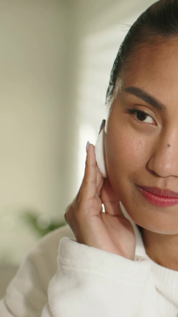 Vertical video: Bringing cotton pad, Asian woman removing eye makeup near blinds wearing white robe
