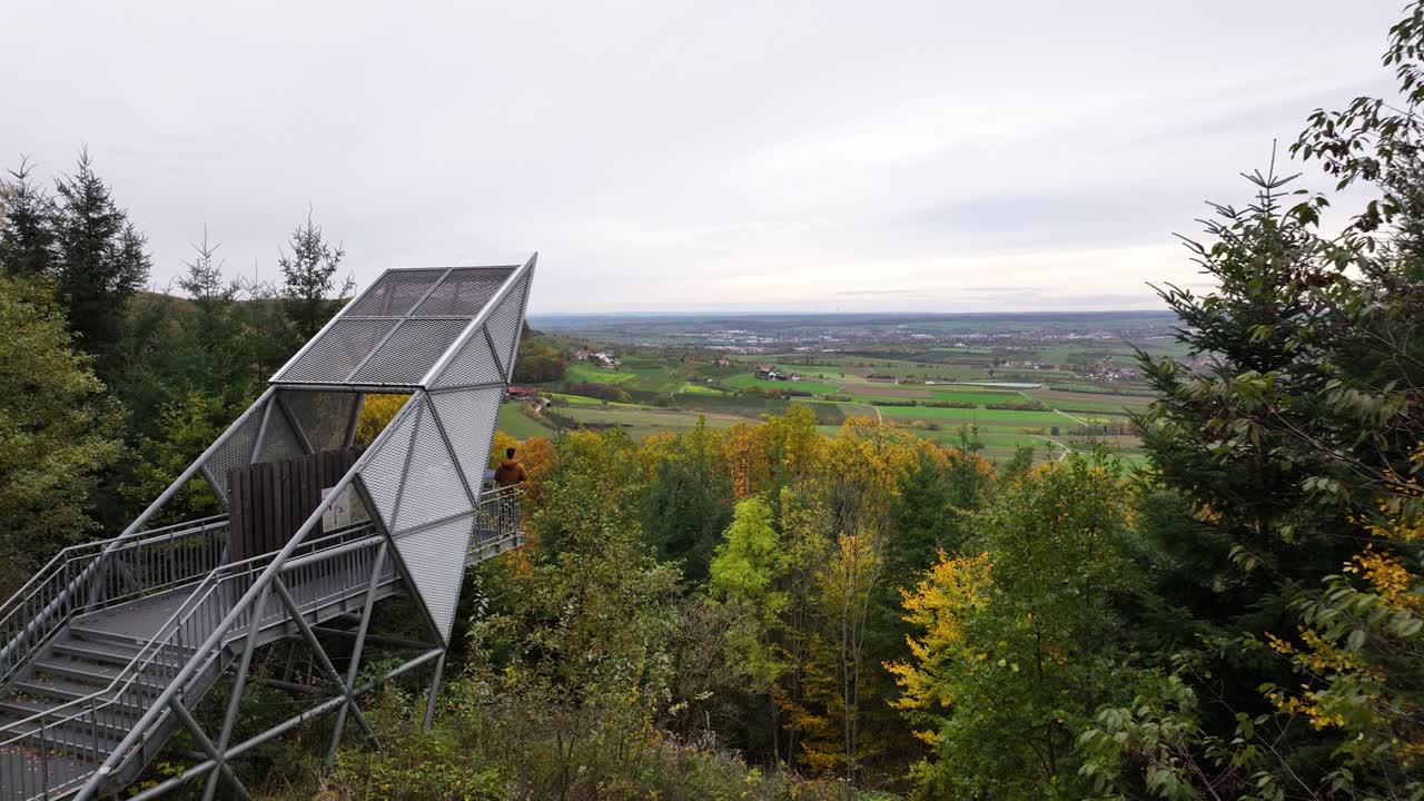 Aerial shot of an observation tower in an colorful autumn forest on a mountain top with a wide and even plateu in the background