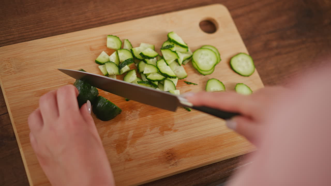Rear view of person holding knife slicing cucumber into small pieces on wooden cutting board, with neat pile of chopped vegetables prepared for healthy meal in kitchen setting