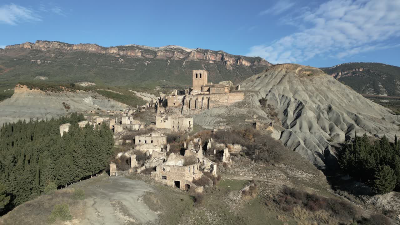 Veduta aerea con movimento a destra che mostra la città fantasma abbandonata di Esco in Aragona, Spagna, con le sue case in pietra in rovina e la chiesa di San Miguel circondata da un paesaggio desolato di calanchi