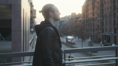 Young-European-Man-with-Glasses,-Beard-and-Man-Bun-Walking-on-Bridge-in-Hamburg,-Germany-with-Cars-in-Background