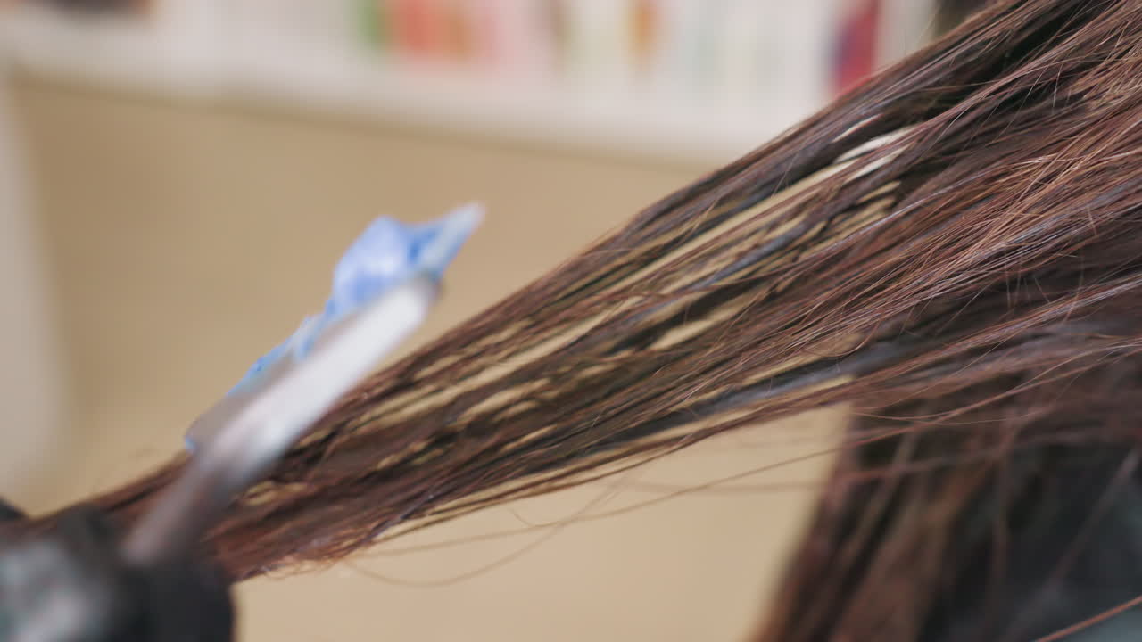 gloved hand applying dye with blue applicator tip to brown hair, careful sectioning with tail comb, targeted pigment placement and professional coloring technique in salon environment