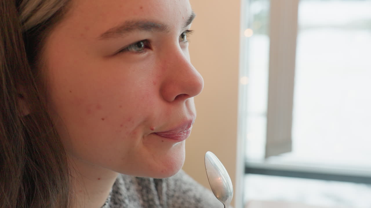 Damsel seated in restaurant tasting her drink from teaspoon blinking in satisfaction with gentle expression cars visible moving across street through large window natural daylight soft atmosphere