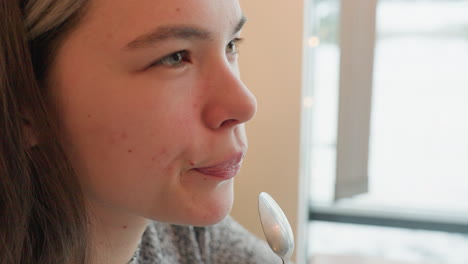 damsel seated in restaurant tasting her drink from teaspoon blinking in satisfaction with gentle expression cars visible moving across street through large window natural daylight soft atmosphere