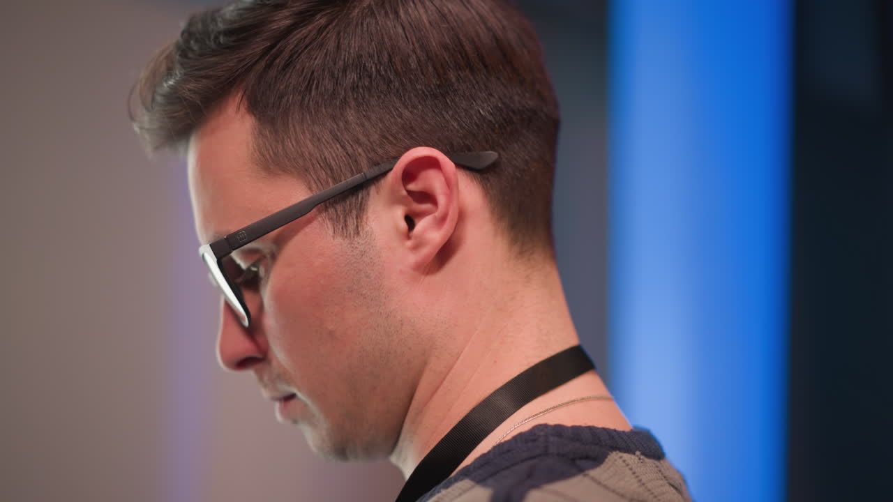 male technician wearing glasses studies monitor reflection under blue LED studio lights with focused expression, head side visible against blurred grid backdrop, conveying concentration during live broadcast prep