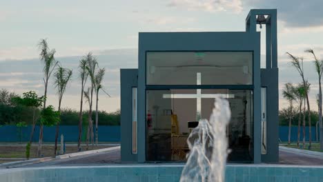 Plano-En-Movimiento-Donde-Se-Ve-En-Primer-Plano-Una-Fuente-De-Agua-Y-Al-Fondo-Una-Pequeña-Capilla-En-Un-Entorno-Natural-Con-Luz-Diurna