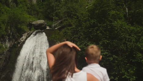 mother adjusts long hair holding toddler son rest by waterfall in rocky park slow motion. young woman shows with boy amazed by water cascade in wood