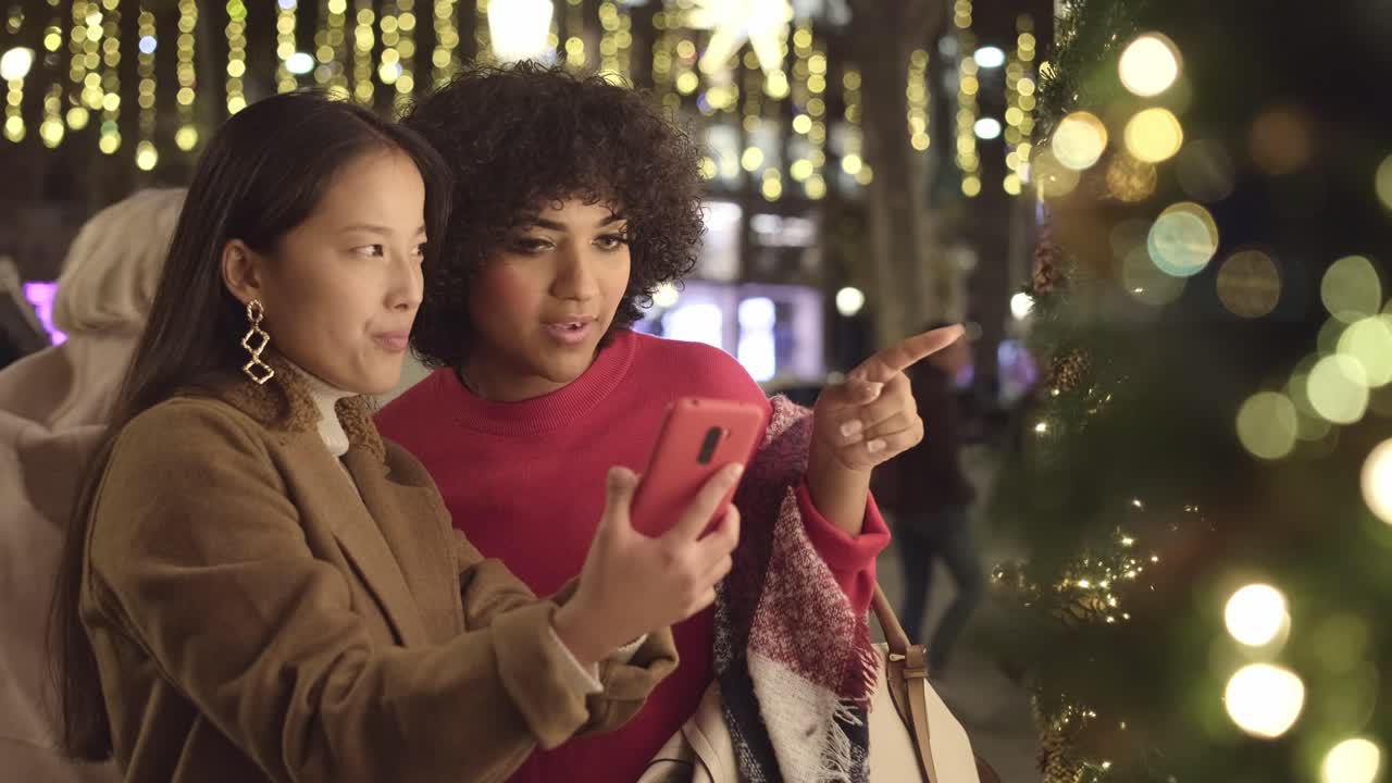 Two friends looking at a smartphone in a city at night with Christmas lights