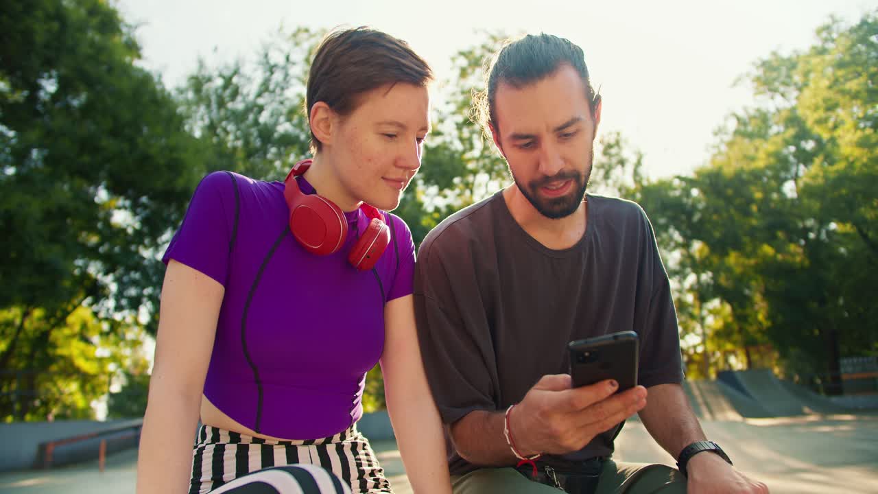 un tipo moreno con una camiseta gris muestra a una chica con un corte de pelo corto en una camiseta púrpura y auriculares rojos algo en su teléfono negro en un skatepark en verano