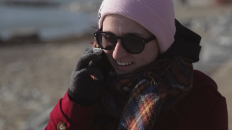 Pretty Woman Wearing Maroon Jacket and Beanie Hat Talking To Phone While Walking On The Street Near Seashore During Sunny Day - Close Up Shot