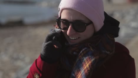 Pretty-Woman-Wearing-Maroon-Jacket-and-Beanie-Hat-Talking-To-Phone-While-Walking-On-The-Street-Near-Seashore-During-Sunny-Day---Close-Up-Shot