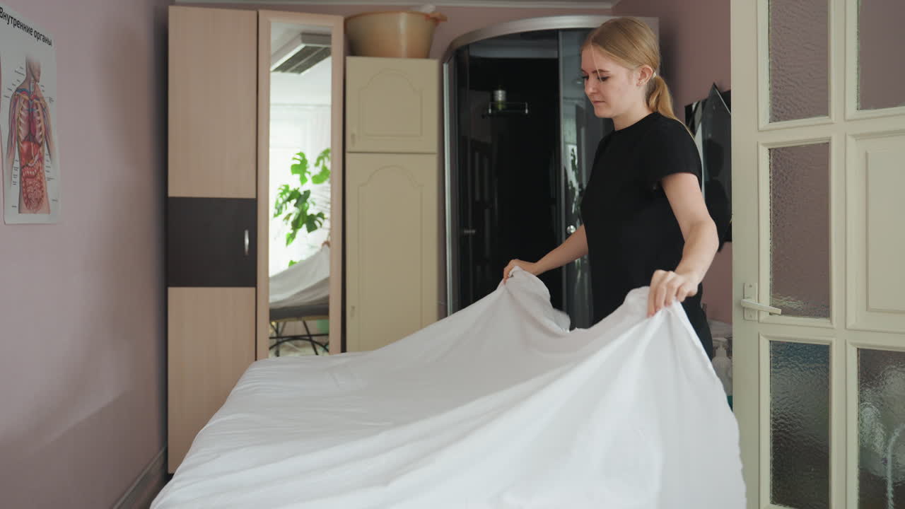 Woman stretches out white bedspread while arranging bed in clean indoor space, her movement subtly mirrored on wall behind her, showcasing preparation, care, and calm atmosphere in softly lit room