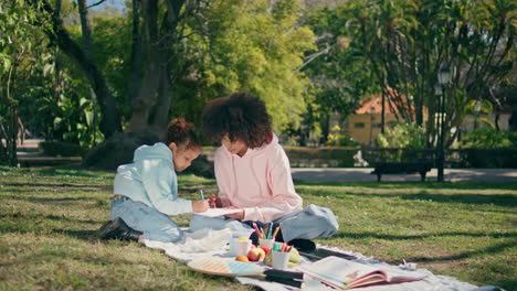 family sitting picnic blanket on sunny green nature. child drawing paper