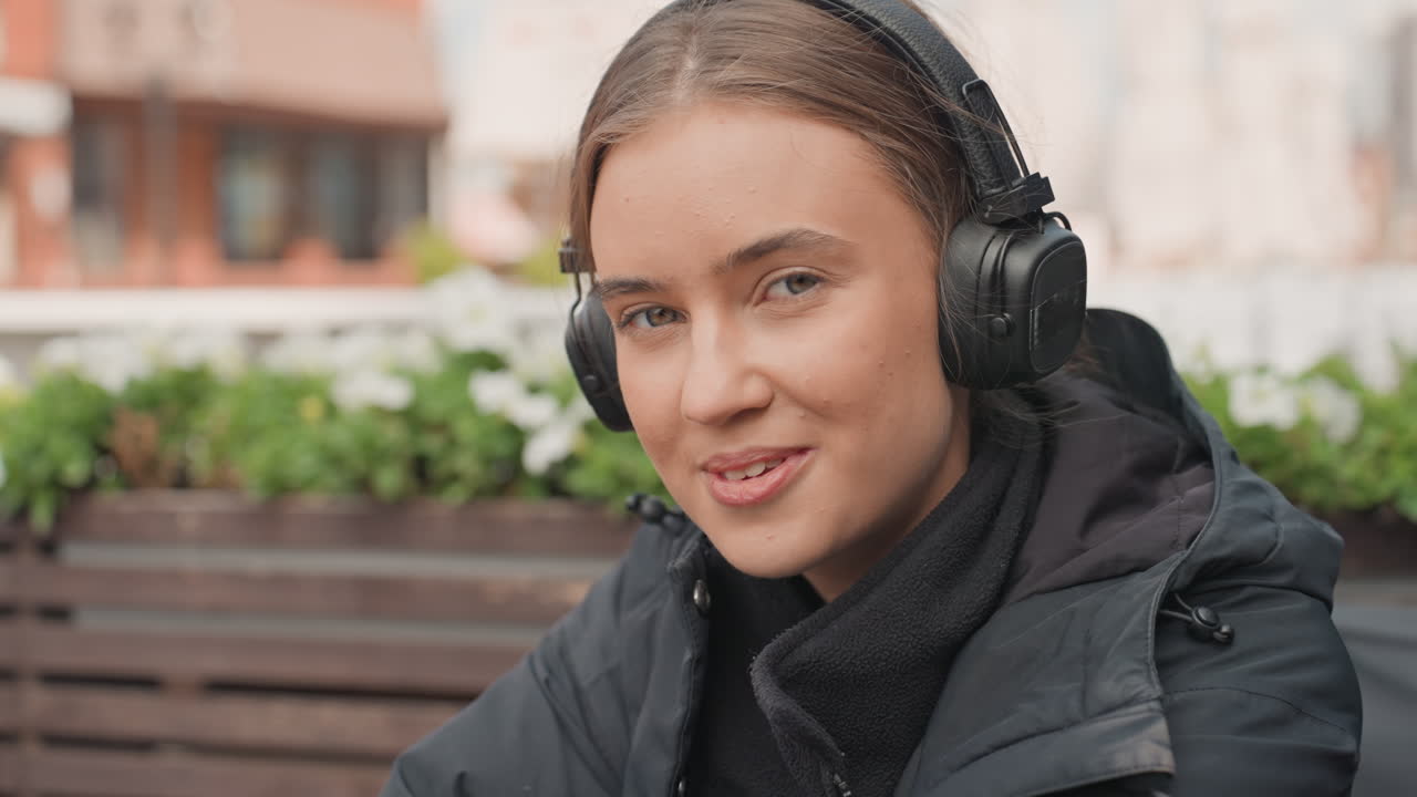 Ung kvinne med hodetelefoner sitter på en trebenk på en utendørs kafé, iført svart jakke og skjerf, smiler mens hun lytter til musikk, diskret øyekontakt med kameraet, murfasader og plantekasser i byen