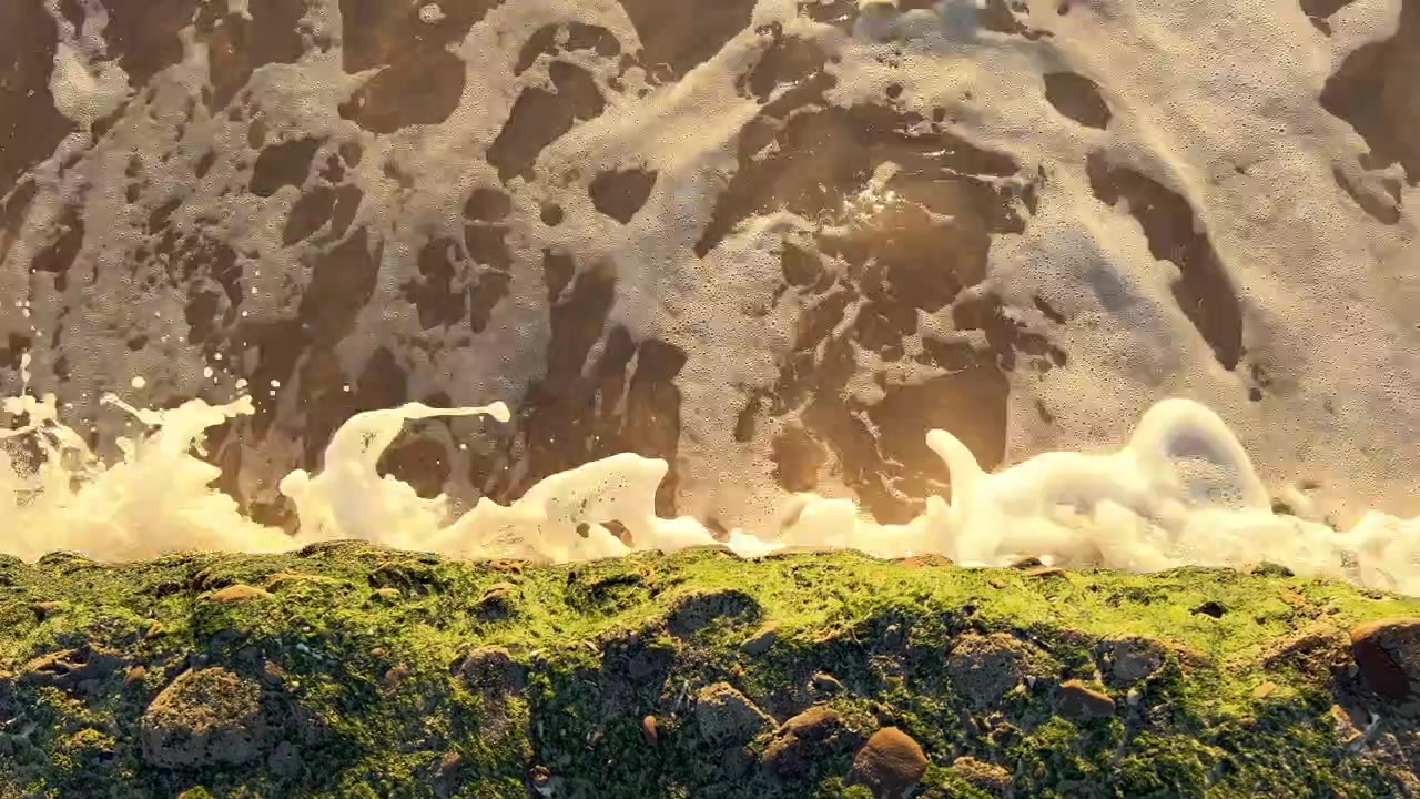 White ocean waves washing over smooth brown sand and algae covered rocks at golden hour in Barbate Spain