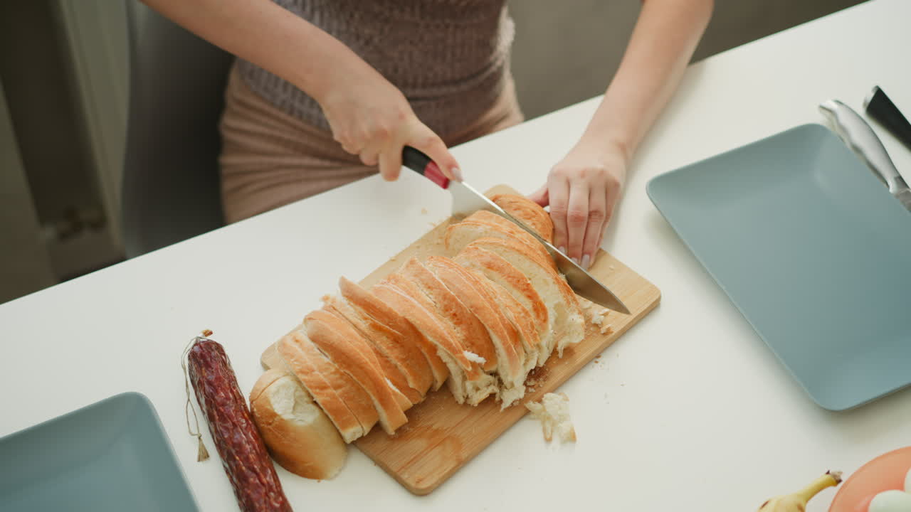 High angle hand view of confectioner slicing cake loaf with knife on white table next to egg banana and bowls in kitchen under bright sunny morning light streaming through window behind