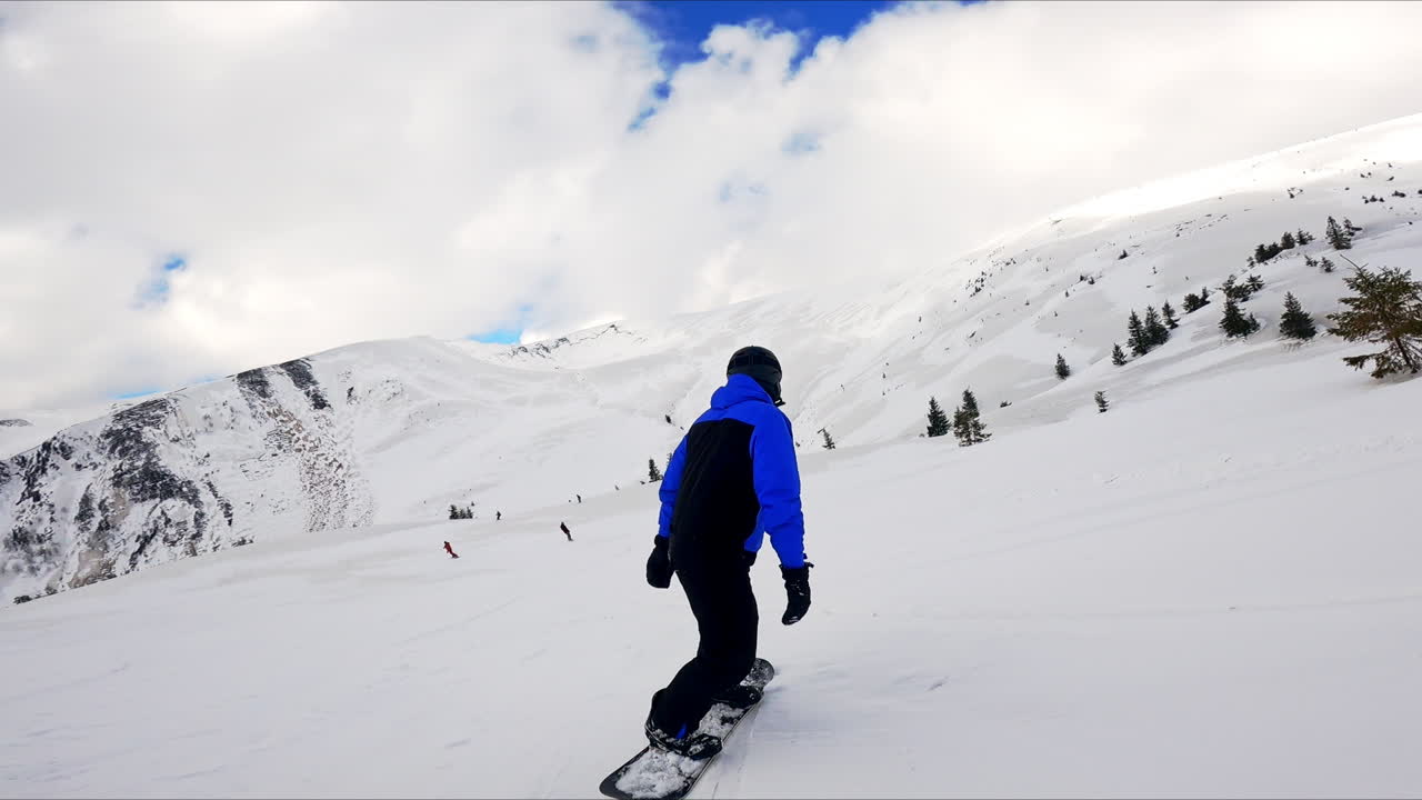 Snowboarder in black and blue sport suit goes down the mountain slope. Amazing snowy scenery under cloudy sky at backdrop.