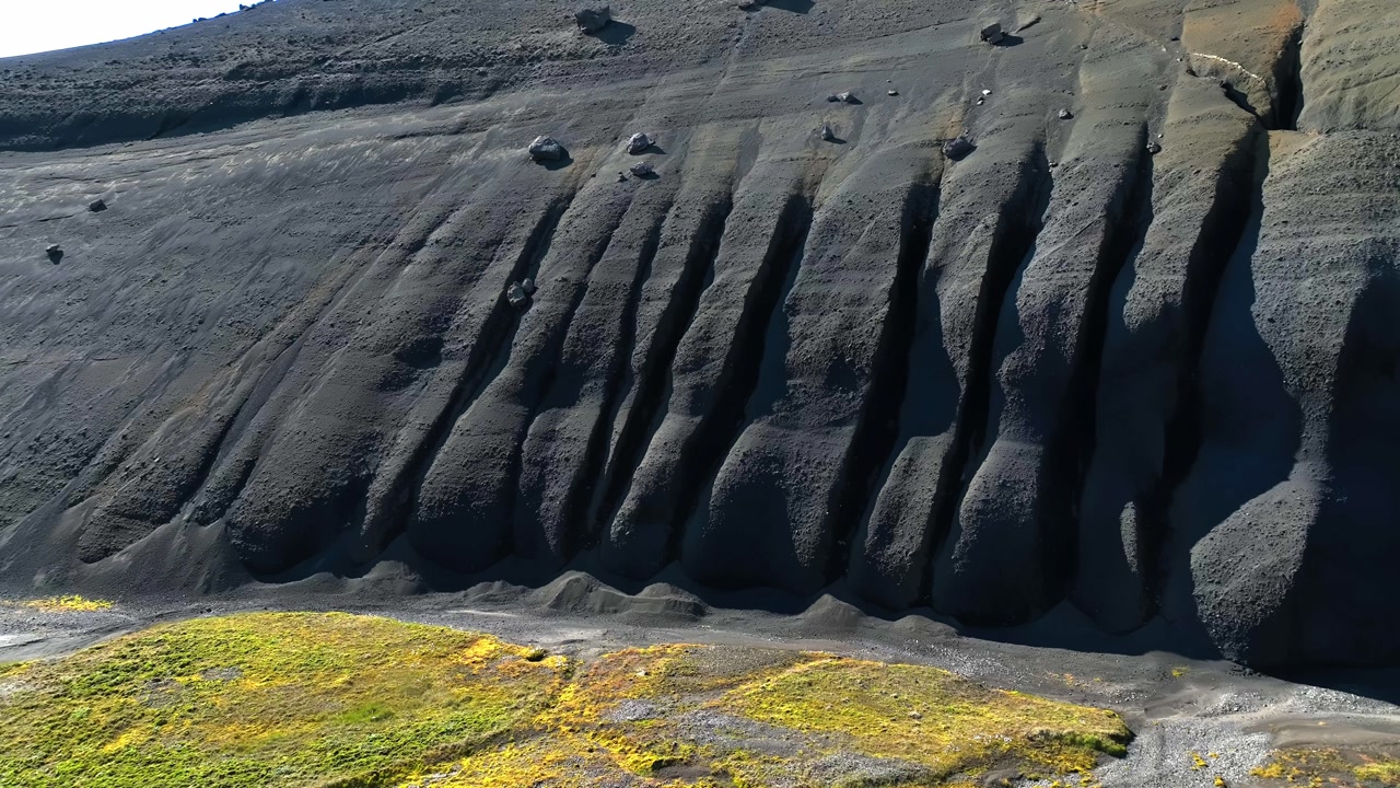 Drone aerial shows a dark volcanic ash cliff with deep vertical grooves and bright moss patches below in Iceland, creating dramatic erosion texture and rugged geology.