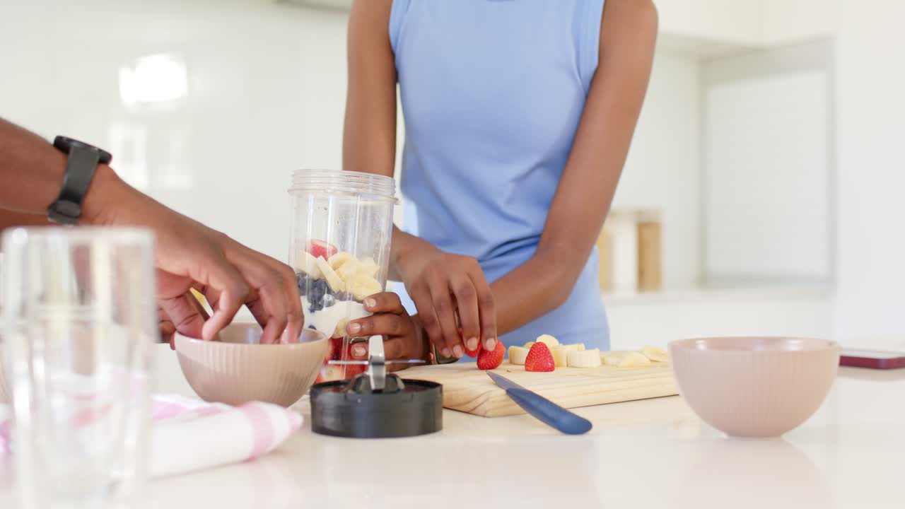 African American couple adding yogurt, steadying blender jar, layering fruit on board for smoothie