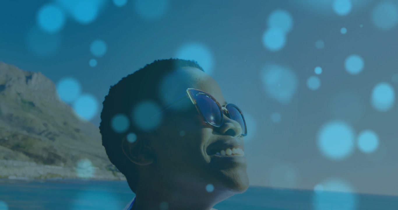 woman wearing reflective shades smiling at seaside, with floating bokeh lights for travel marketing