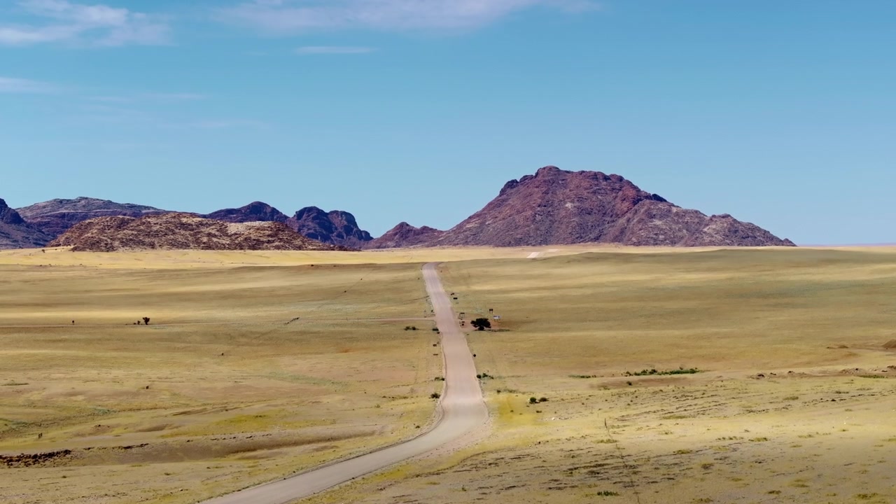 Vista aérea de una sinuosa carretera desértica que conduce a una prominente montaña roja a través de las doradas llanuras de Namibia. Llanuras de hierba dorada se extienden hacia impresionantes montañas de granito rojo bajo un brillante cielo azul desértico.