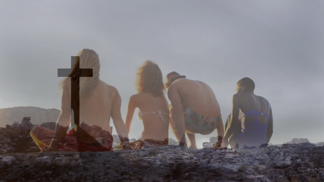Sitting on rocky landscape, diverse group watching silhouetted cross at sunset