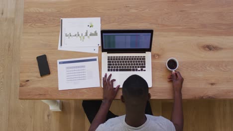 Overhead-view-of-african-american-drinking-coffee-and-using-laptop-while-working-from-home