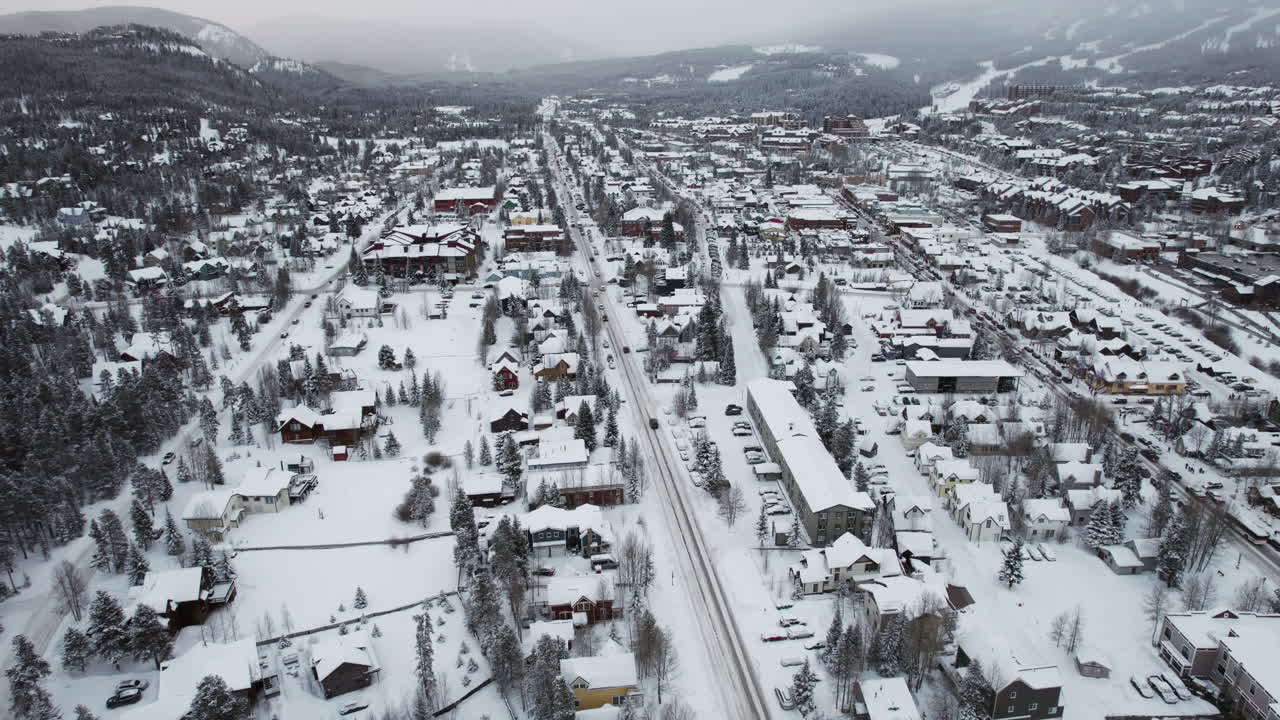 A wide rising aerial of Breckenridge, Colorado, peaceful, beautiful and snow-covered in winter, with cars carrying skis and snowboards into the popular mountain town and world famous resort.