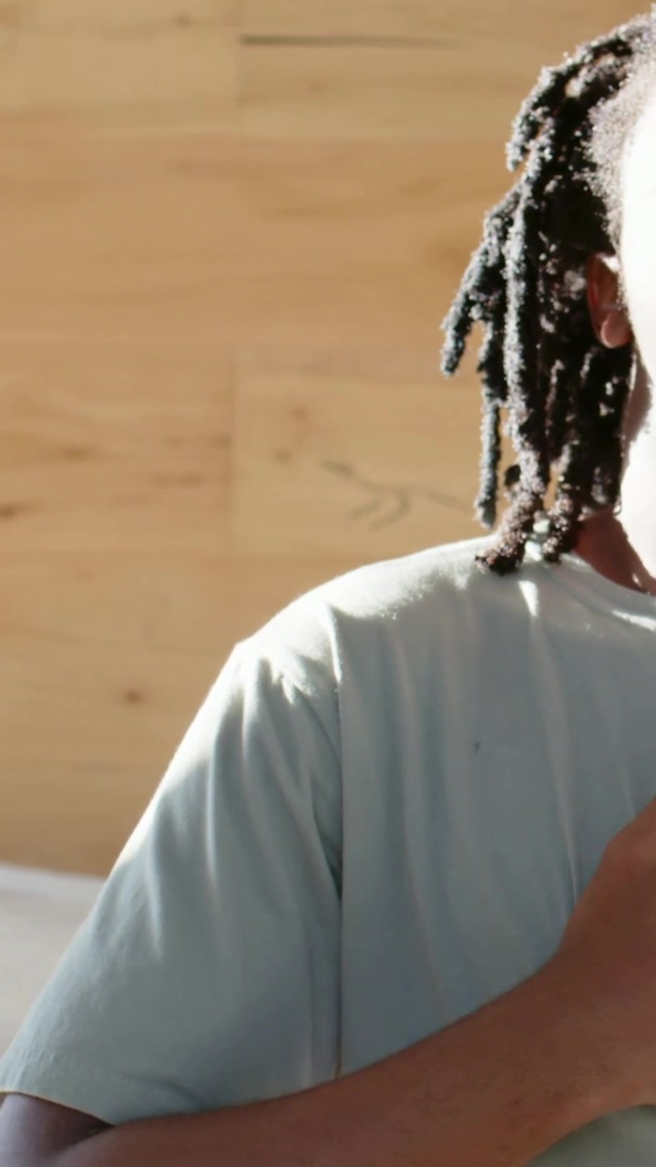 Video verticale: Uomo seduto con i dreadlock, indossa una maglietta chiara, tiene un dispositivo sul letto, con spazio copia.