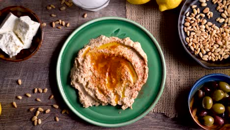 wide sliding to the left showing a flat lay plate of hummus with feta, olives, glass of ouzo, lemons, pine nuts on a rustic wood table