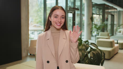 happy young caucasian businesswoman looking at camera and waves hand hello in office hotel lobby