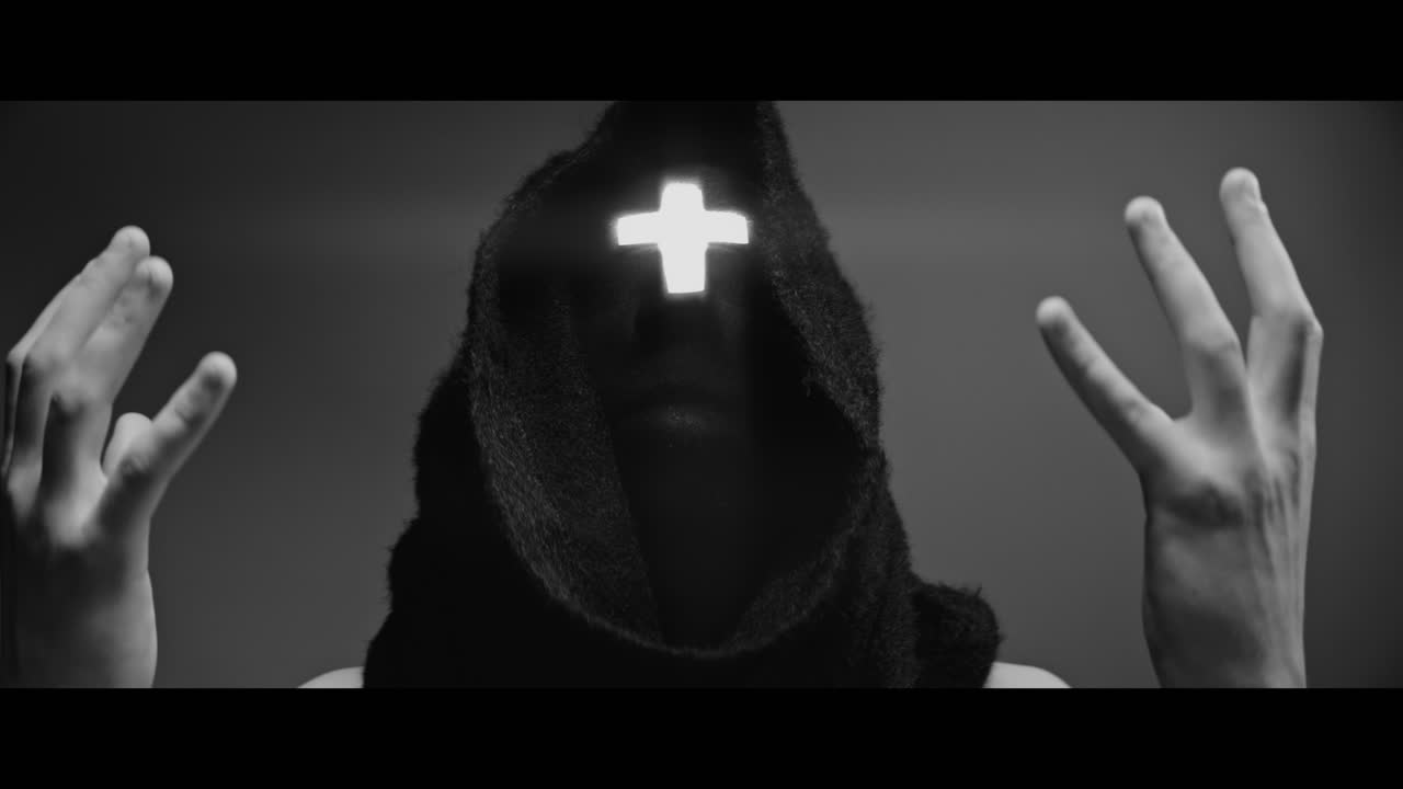 Masked and hooded man with cross of light glowing on his face looks in the camera