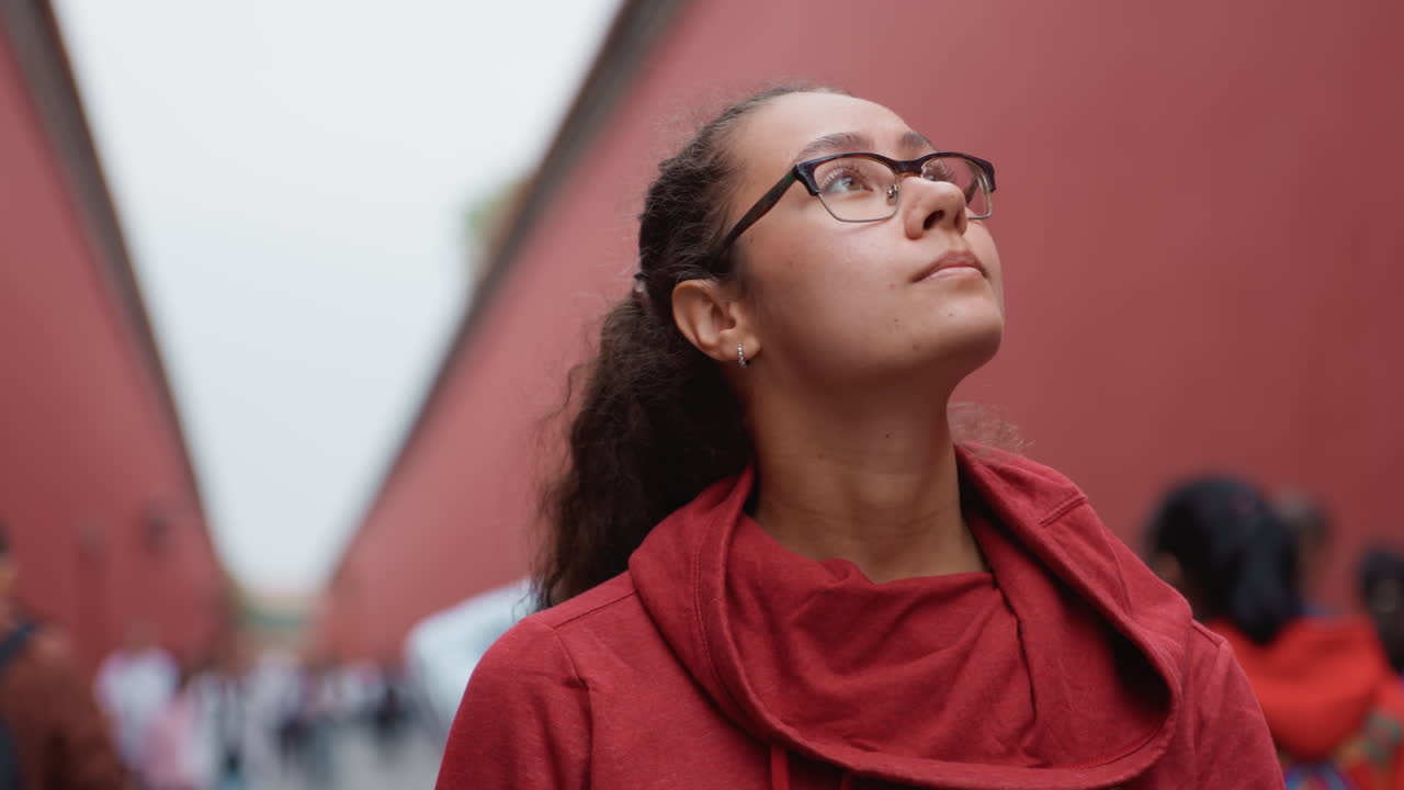 Young woman with glasses looking upward in narrow red alley, wearing red scarf, soft overcast sky, blurred crowd background, contemplative expression, urban tourist exploring architecture, warm color