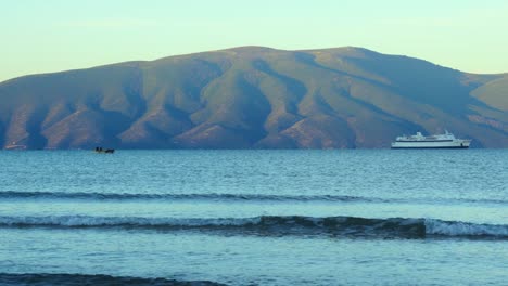 Fishermen-at-sunrise-with-fishing-boat-floating-on-sea-water-near-shore,-white-ferry-approaching-slowly-toward-port