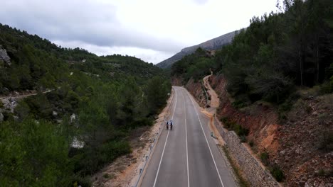 panorámica de imágenes de drones de tres ciclistas del equipo recorriendo una pintoresca carretera española, a través de un valle con tierra roja expuesta, un desfiladero de río y frondosos árboles verdes que bordean la carretera