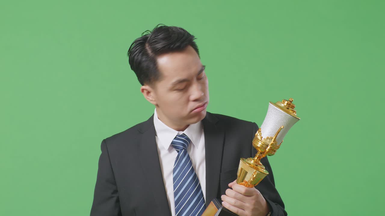 화가 난 아시안 사업가, 슈트와 타이를 입고, 금메달을 달고, 트로피를 쳐다보고, 머리를 흔들고, 불만을 품고, 스튜디오의 녹색 스크린 배경에서 첫 번째 우승자로 승리합니다.