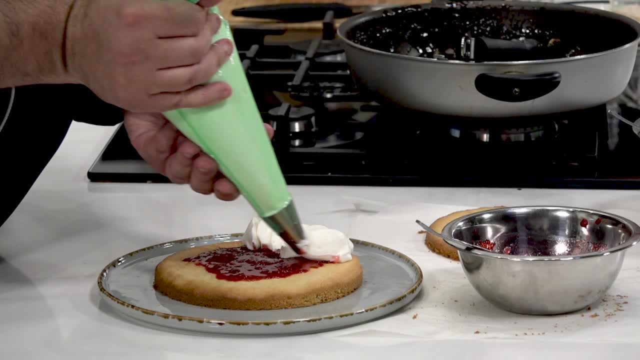 Baker skillfully pipes fresh jam and whipped cream onto a cake layer in a warm and inviting kitchen. Delicious treats are in the making, showcasing culinary expertise