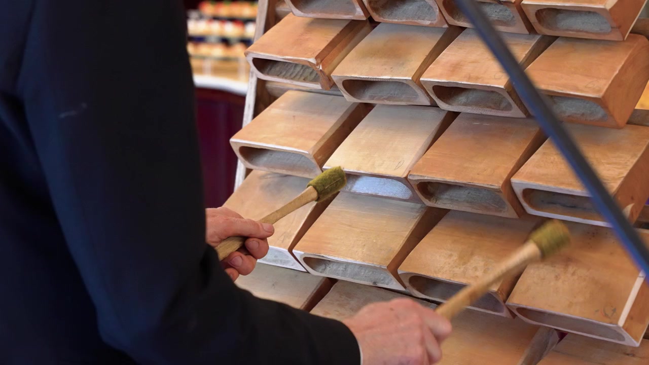Un percussionista si esibisce su un tamburo a lingua in legno unico, colpendo pannelli testurizzati con bacchette al chiuso. L'illuminazione calda, la composizione ravvicinata e il movimento ritmico creano una scena artistica e musicale.