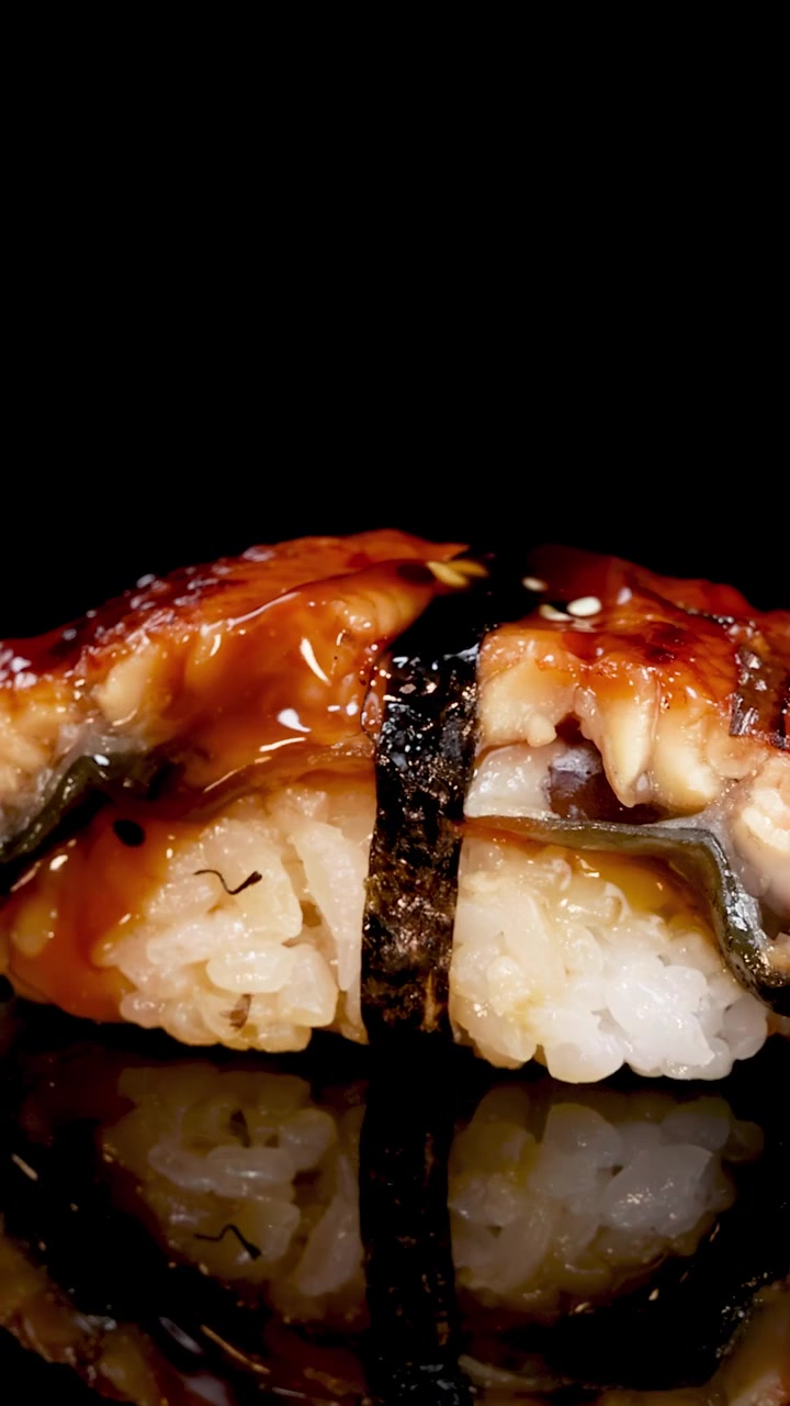 Un pezzo di sushi nigiri unagi ruota dolcemente sotto un'illuminazione drammatica, mettendo in risalto l'anguilla lucida, il riso e il nori su una superficie nera riflettente