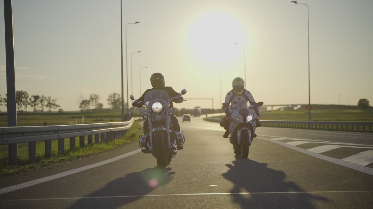 dos motociclistas montando diferentes tipos de motocicletas golpeándose las manos mientras viajaban uno al lado del otro en una carretera vacía durante la puesta de sol