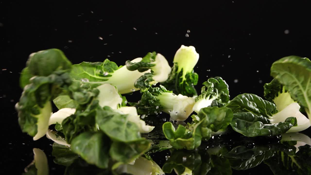 Bok choy leaves drop onto a glossy black surface, creating a dynamic visual with vibrant greens and smooth reflections