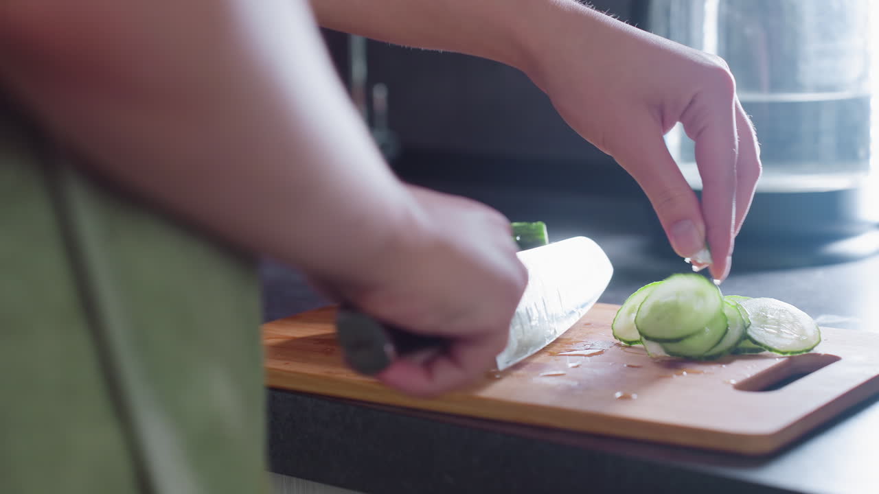 Close-up of lady's hand cutting cucumber into smaller pieces on a wooden cutting board, focus on precision, healthy food preparation in bright kitchen environment