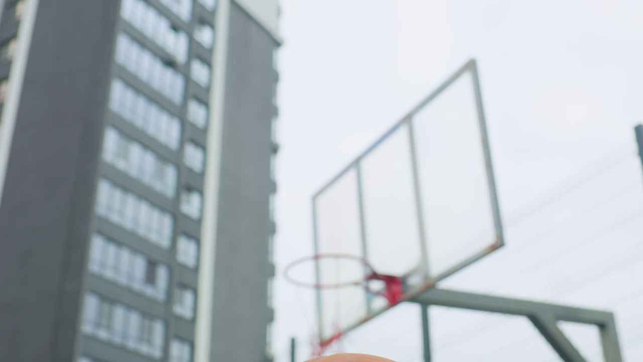 Primo piano di un canestro da basket. Un giovane atleta in equilibrio con un dito tiene una palla sulla punta del dito mentre mira al canestro su un campo pensile, con grattacieli sullo sfondo, luce fresca e nuvolosa, sessione di allenamento concentrata.