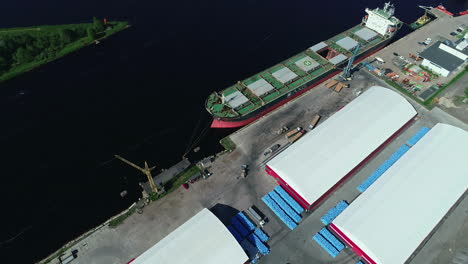 aerial view of cargo ship docked at harbor with adjacent warehouses and lush riverbank on sunny day