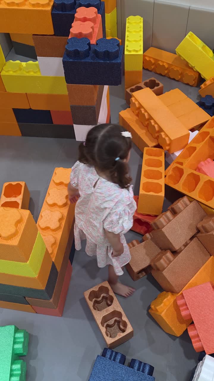 A young girl develops creativity and motor skills while playing with giant, colorful foam building blocks, constructing a tower at an indoor playground or kids' cafe.