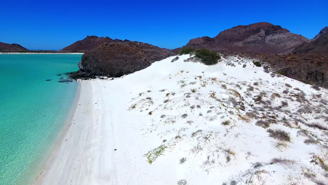 Forward Flying View of Women Walking on a White Sand Beach with Blue Shades of Water and Mountain Background