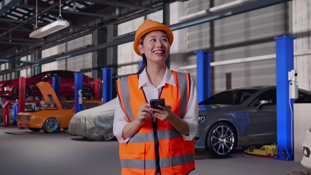 Ingegnere donna asiatica con casco di sicurezza che utilizza lo smartphone e si guarda intorno mentre si trova in un'autofficina