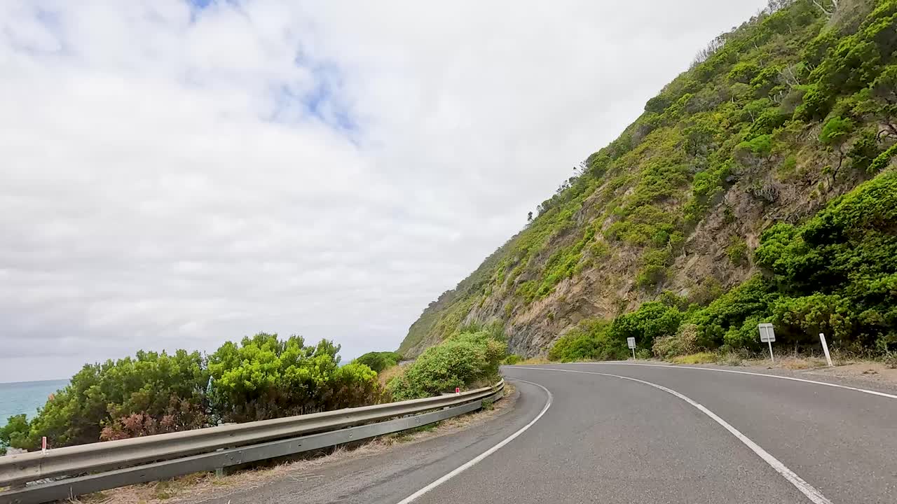 A car travels along the picturesque Great Ocean Road, flanked by cliffs and ocean under a partly cloudy sky