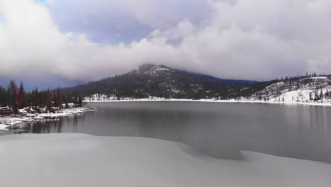 aerial pullback over partially frozen lake and snow-covered forested shores with beautiful snowy mountain touched by clouds in blue sky in background